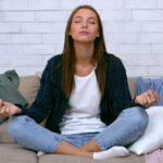 a-young-woman-is-sitting-cross-legged-on-a-couch-eyes-closed-in-meditation-surrounded-by-scattered-clothes-in-a-cozy-living-room-filled-with-soft-lighting-video